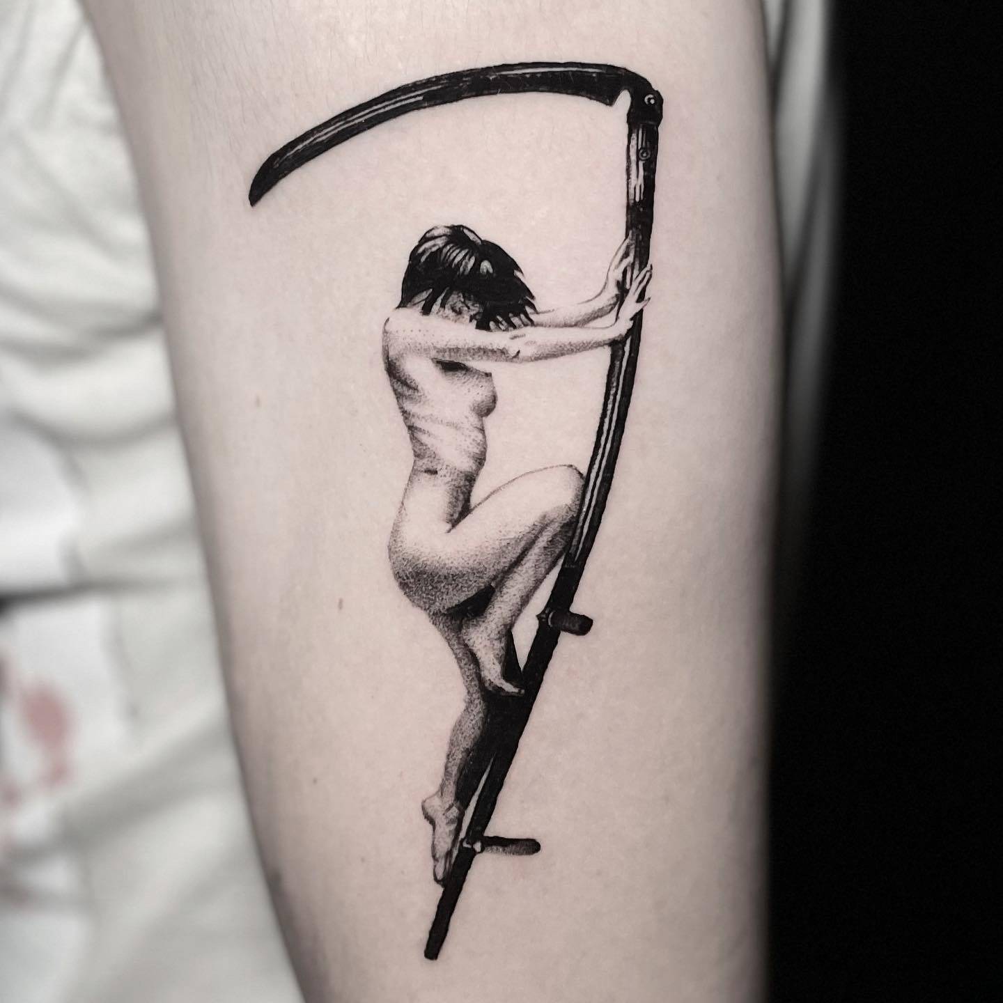 Black and grey tattoo of a seated figure balanced on the blade of a scythe, on the upper arm.
