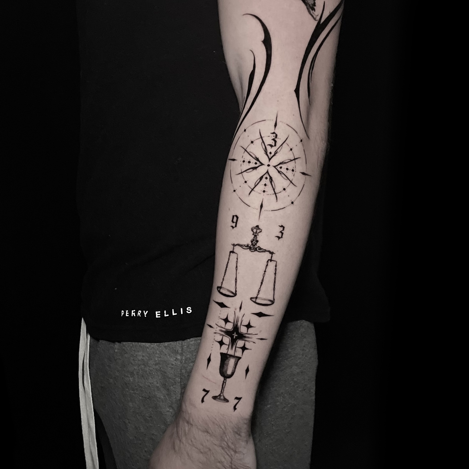 Fine black ornamental tattoo of a compass star, scales and goblet with numerals on the forearm.