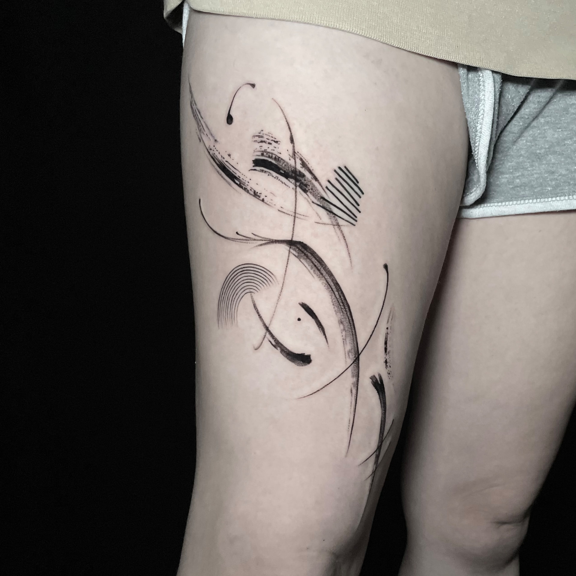 Abstract blackwork tattoo with sweeping lines, brush-like marks and fine arcs on the thigh.