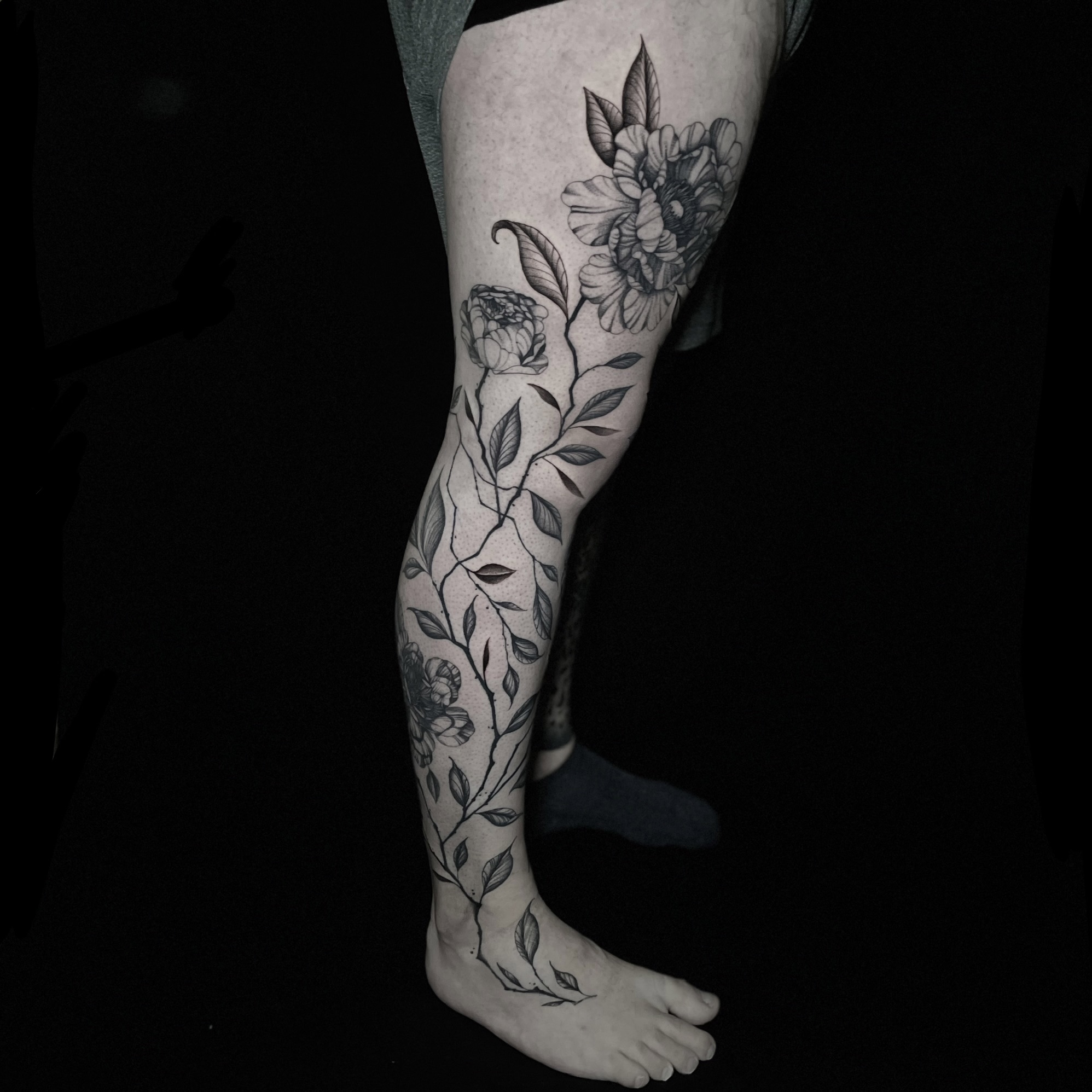 Black and grey botanical tattoo of peonies and branches flowing down the full length of the leg.