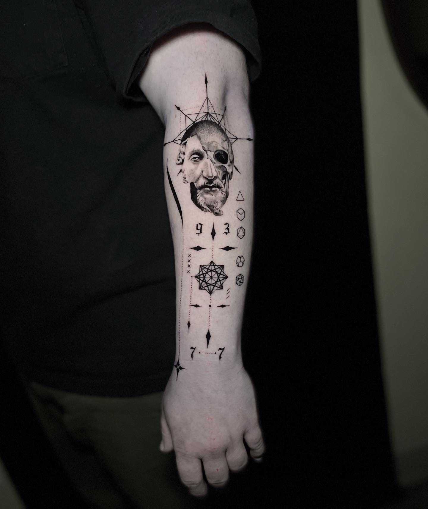 Black and grey forearm tattoo of a split portrait and skull with geometric symbols and numerals extending to the hand.