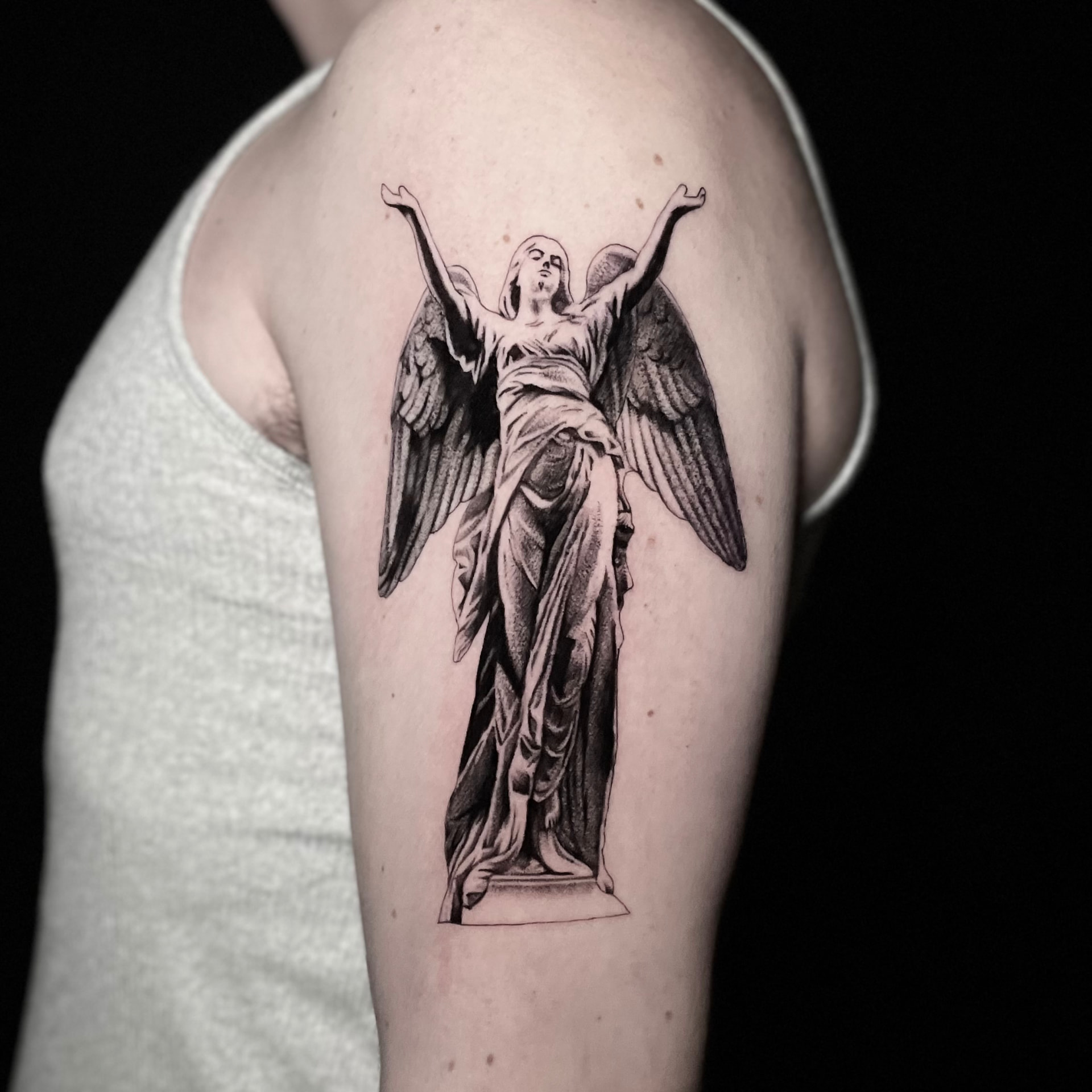 Black and grey angel statue tattoo with outstretched arms on the upper arm.