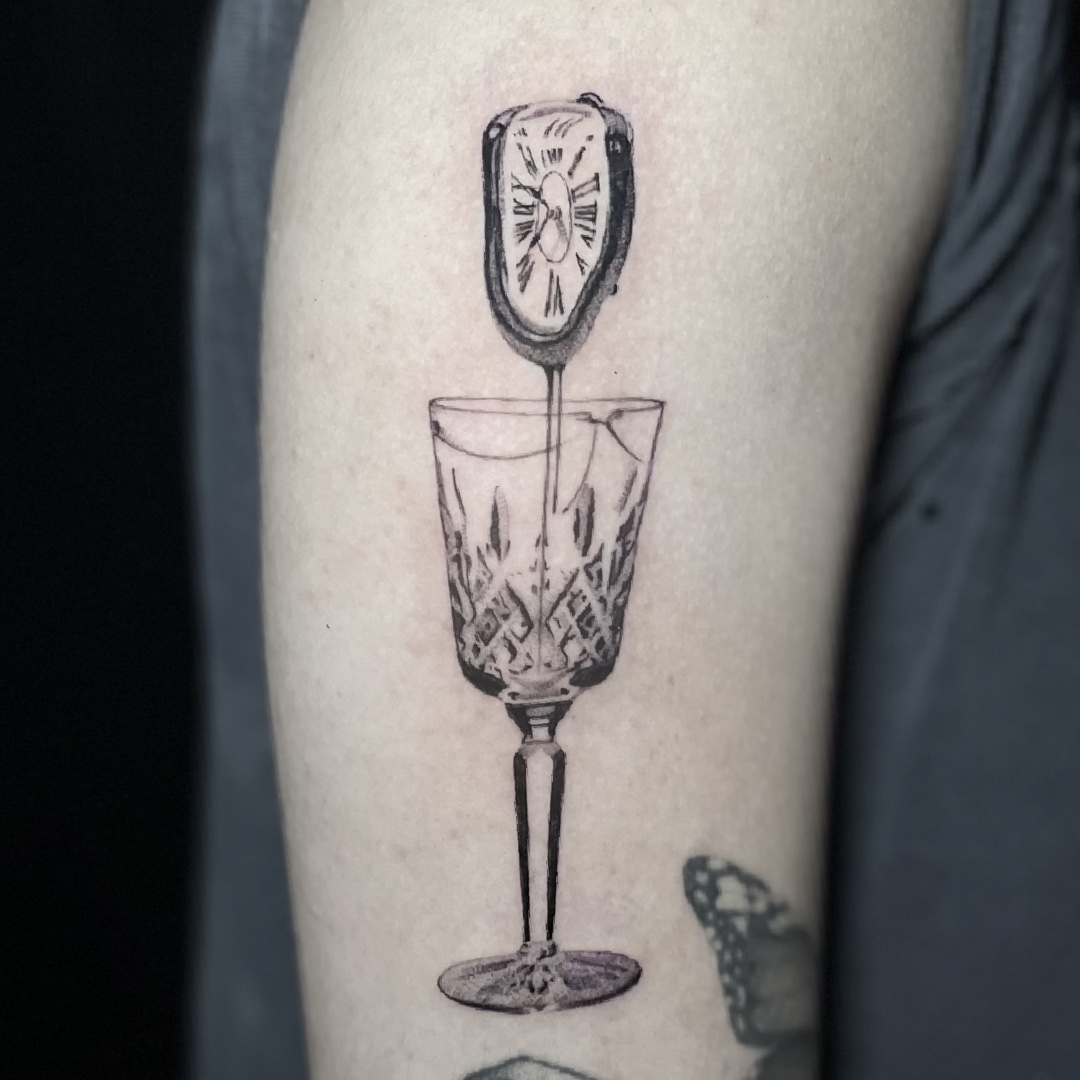 Black and grey tattoo of a melting clock dripping into a crystal goblet on the forearm.