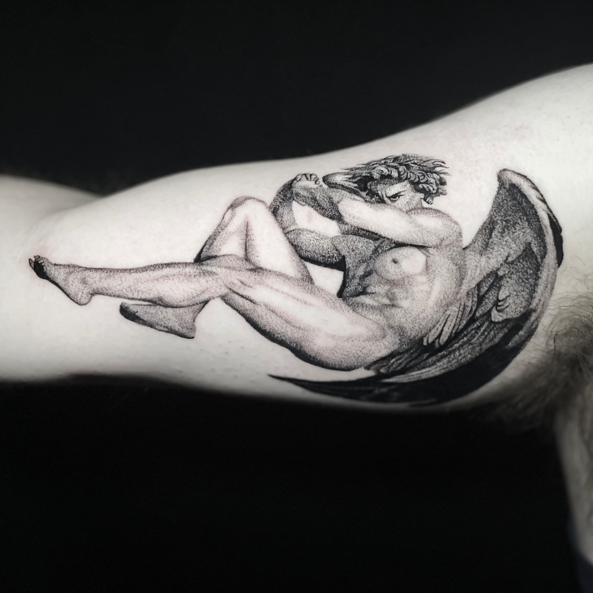 Black-and-grey fallen angel statue tattoo on the inner upper arm with stipple shading.