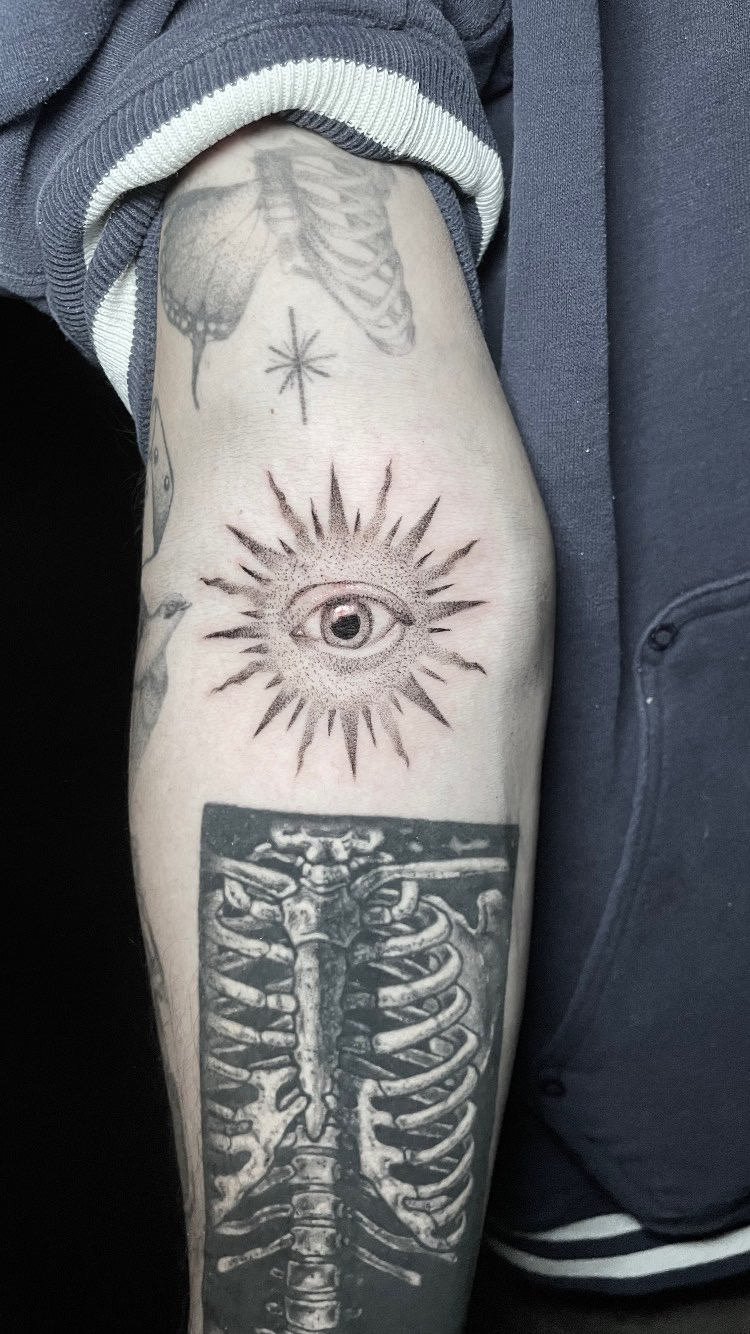 Fine line eye tattoo surrounded by stippled rays on the upper arm.