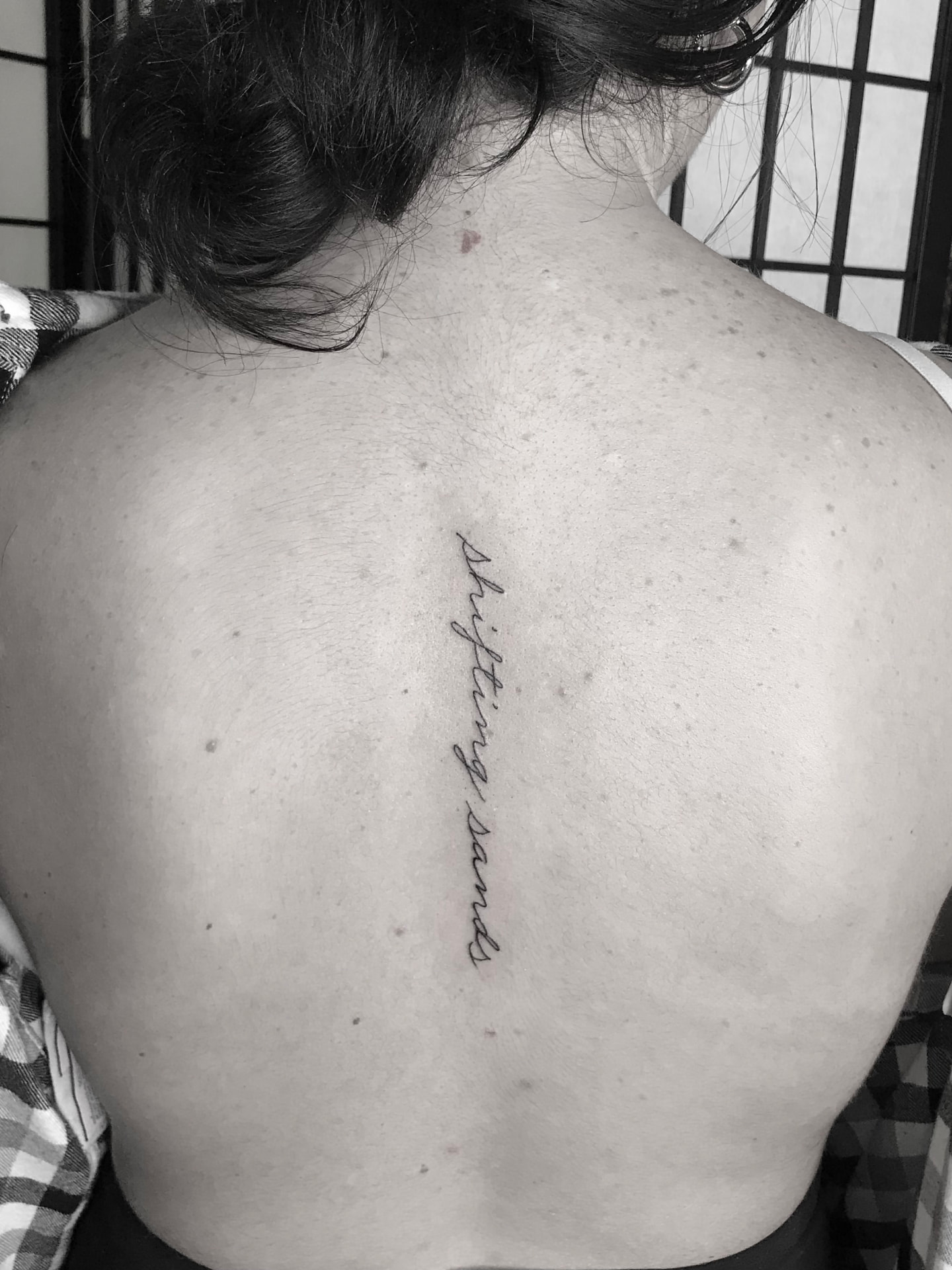 Fine line script tattoo reading shifting sands, placed vertically on the upper back.