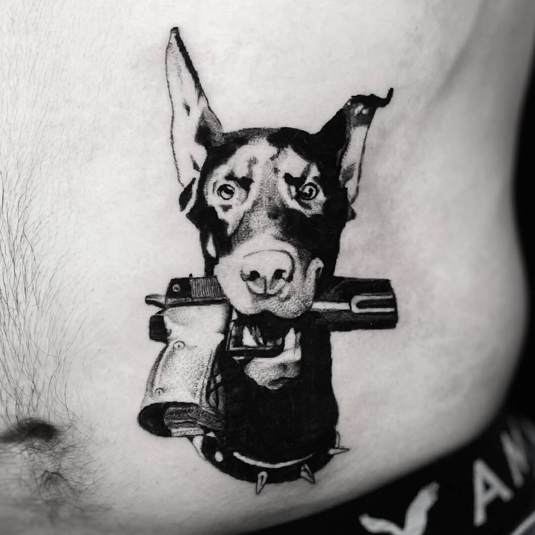 Black and grey realism tattoo of a doberman holding a pistol, tattooed on the ribs.
