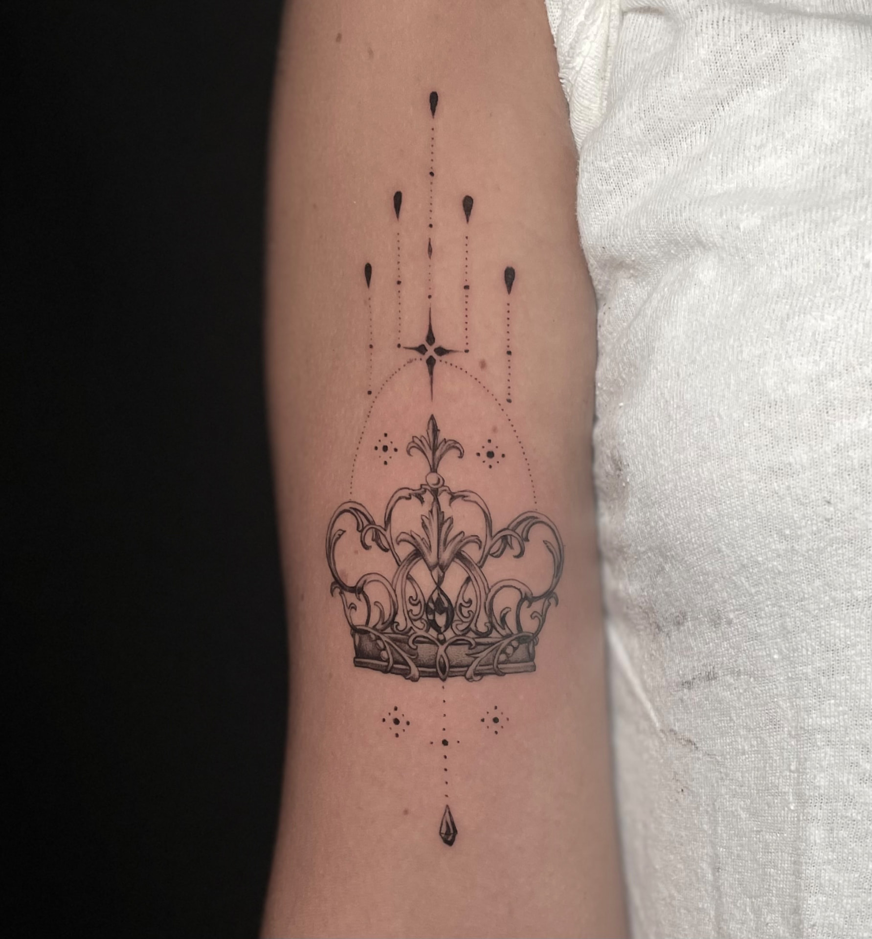 Fine line ornamental crown tattoo with stars, dots and teardrop details on the upper arm.