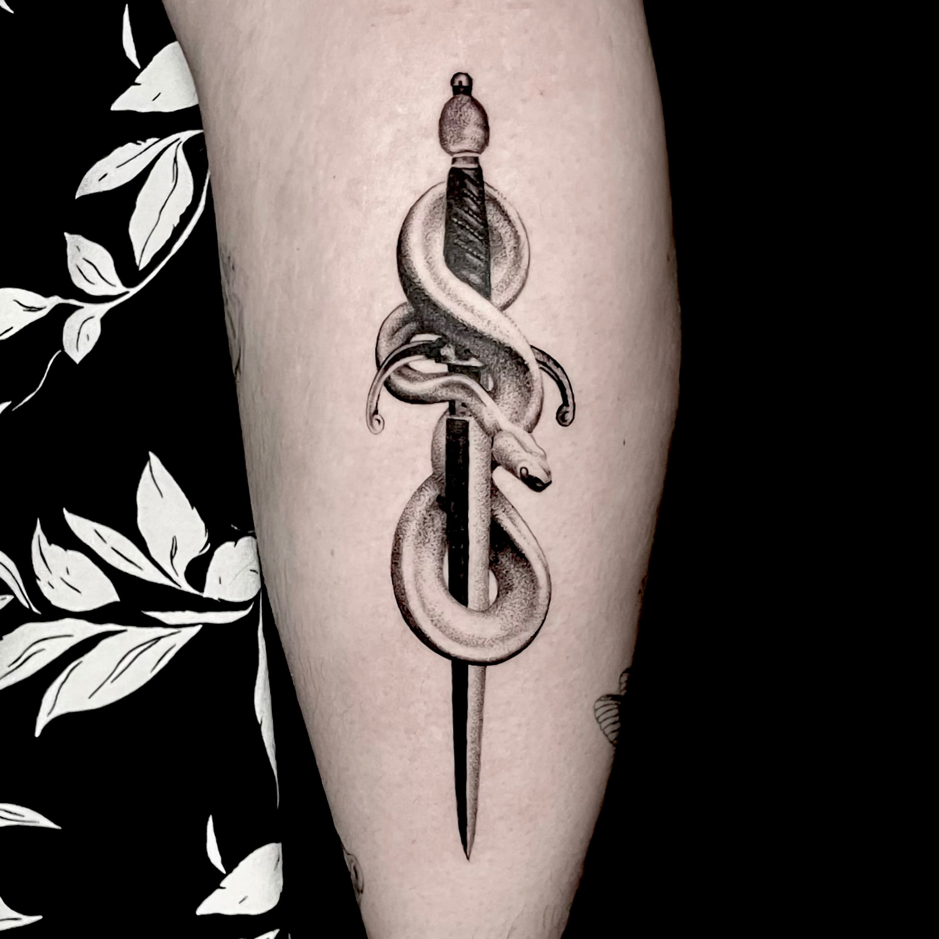 Black and grey tattoo of a snake coiled around a dagger on the forearm.