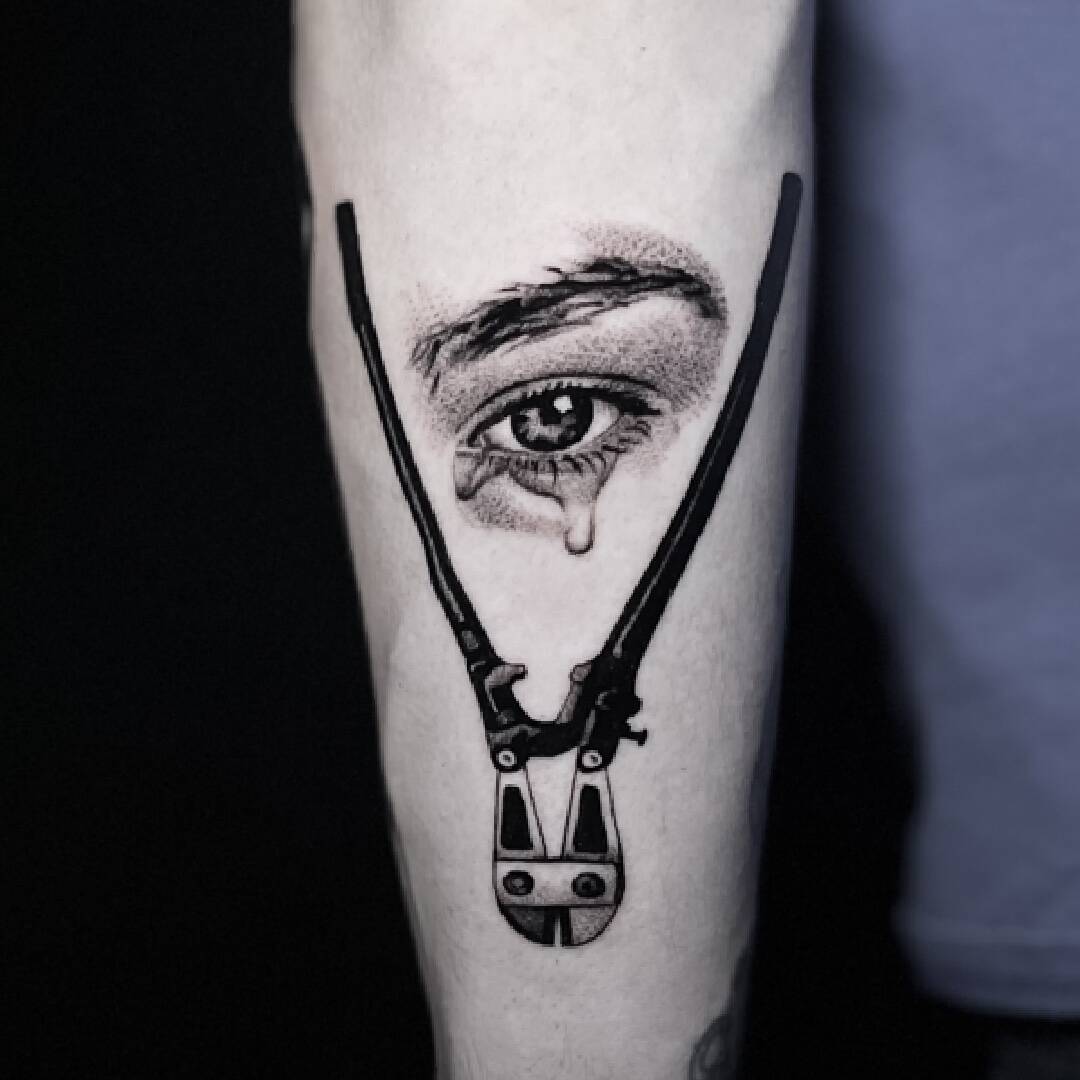 Tattoo of a realistic crying eye held open by a black mechanical clamp, on the forearm.
