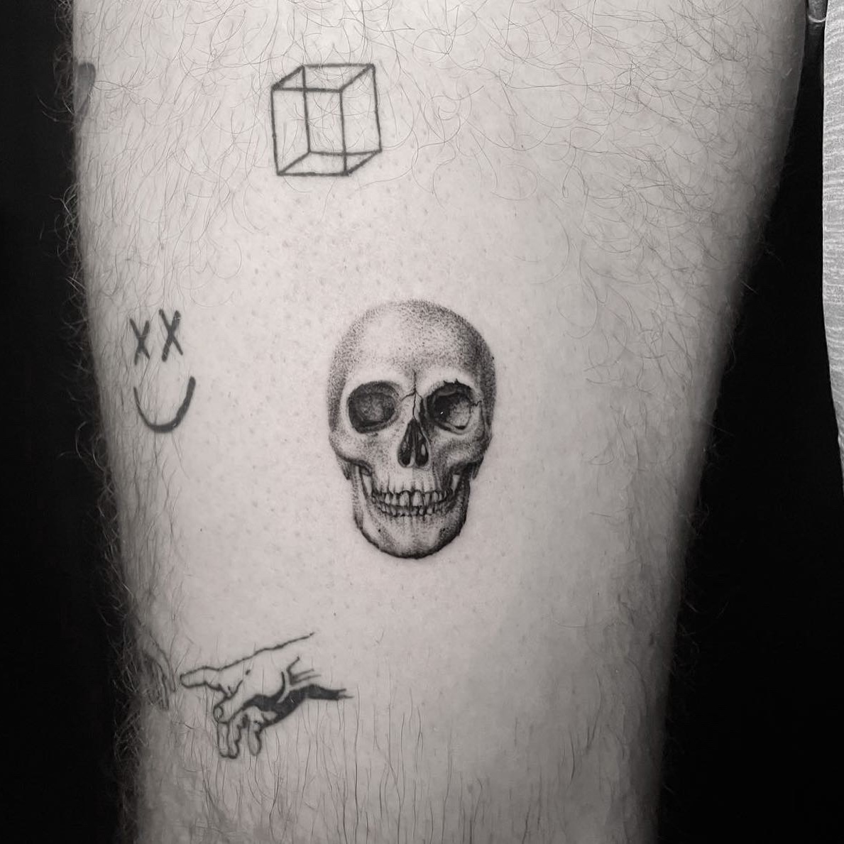 Small black and grey realism skull tattoo surrounded by tiny linework symbols on the thigh.
